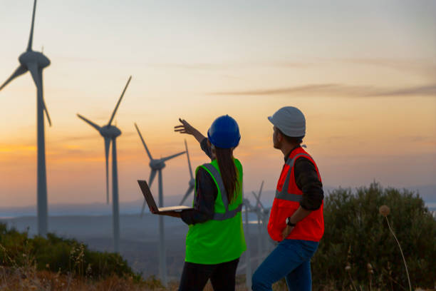Young electrical engineer woman and business man standing in front of wind turbines checking and working about technical problems and writes the results of measurements with laptop pc in wind power plant electric energy station.